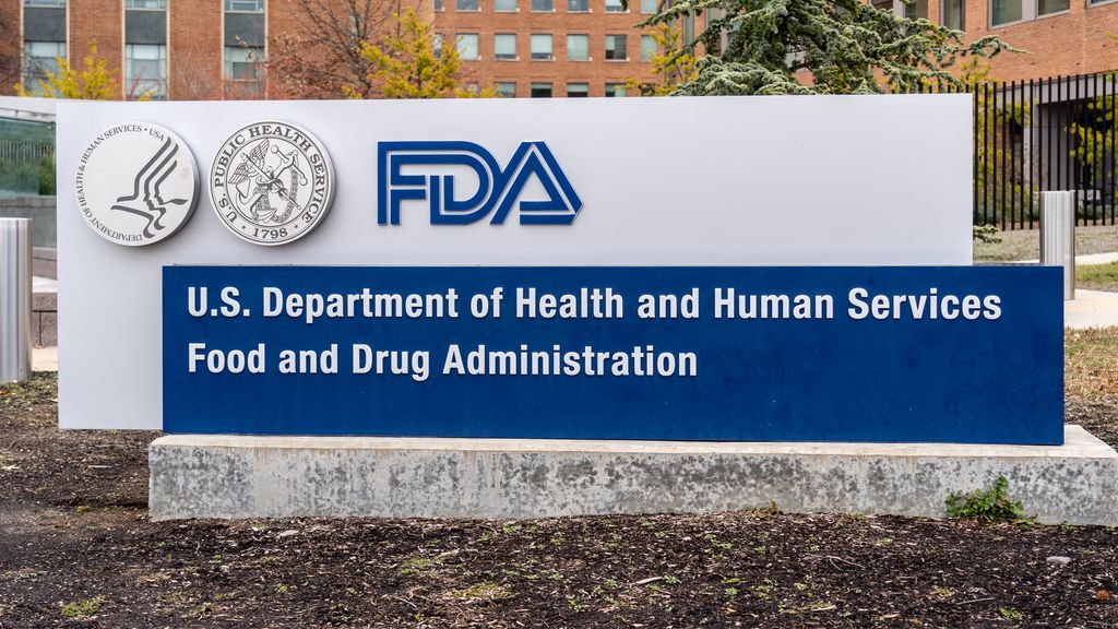 The image contains a stone sign reading: FDA, U.S. Department of Health and Human Services Food and Drug Administration. There is a corporate building in the background and soil ground in front of the sign.